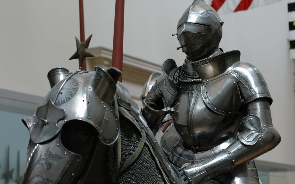 This photograph shows four mounted Knights at the Arms and Armor Department at the Metropolitan Museum of Art in New York. It also shows a collection of various pieces of arms and armor.