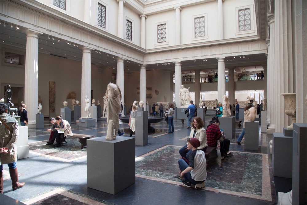 This photograph shows a large gallery enclosed by a colonnade filled with stone sculptures from ancient Rome and Greece. The gallery is at the Metropolitan Museum of Art in New York City.