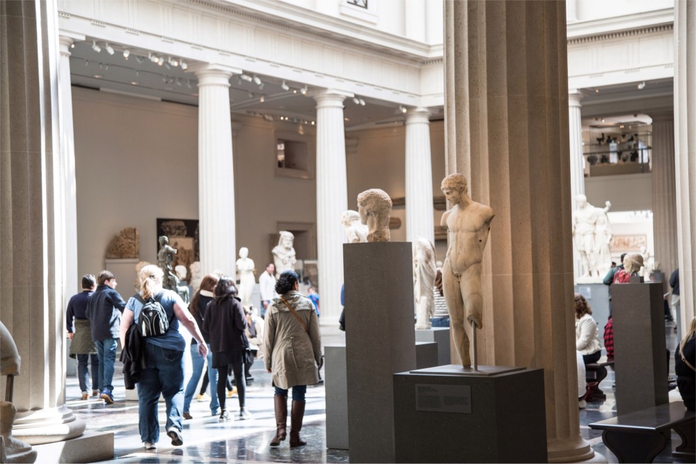 This photograph shows a large gallery enclosed by a colonnade filled with stone sculptures from ancient Rome and Greece. The gallery is at the Metropolitan Museum of Art in New York City.